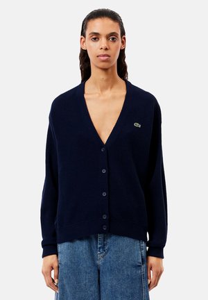 Cardigan a maglia navy con scollo a V profondo, apertura con bottoni e polsini a costine. Presenta un piccolo logo verde sul lato sinistro del petto. Texture morbida.