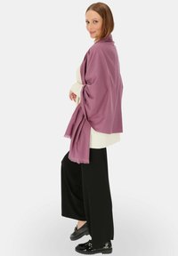 Jasnofioletowa chusta opada na białym swetrze, połączona z czarnymi spodniami o szerokich nogawkach i lśniącymi czarnymi loafersami. Charakteryzuje się frędzlami na krawędziach.