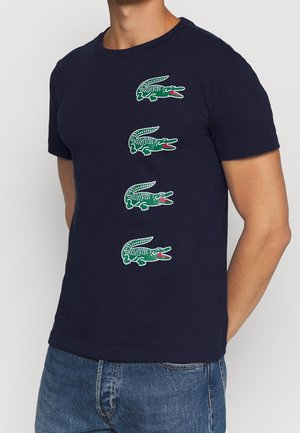 Homme portant un t-shirt bleu marine avec quatre logos verts d'alligator alignés verticalement sur le devant et un jean bleu.