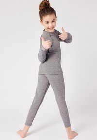 Maglia grigia a maniche lunghe e leggings aderenti in morbido materiale, caratterizzati da un design semplice e un logo sulla manica. Modella che fa segno di approvazione.
