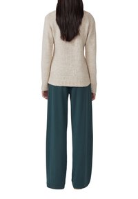 Persona con lunghi capelli scuri che indossa un maglione di maglia beige e pantaloni verdi scuri larghi, in piedi con la schiena rivolta verso la fotocamera su uno sfondo bianco.