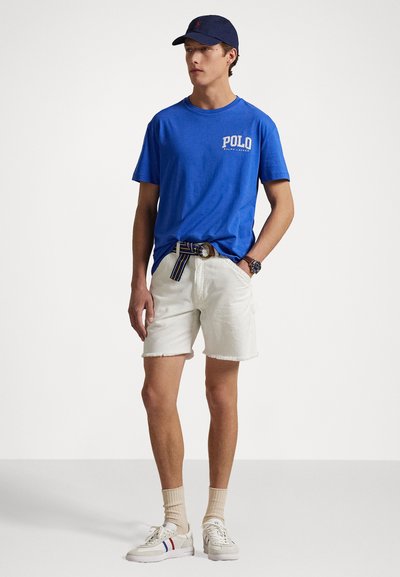 Blå bomullströja med "POLO"-logotyp, vita denimshorts med fransar, randigt bälte, beige strumpor och vita sneakers med färgaccenter.