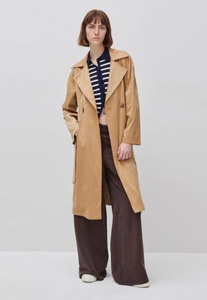Donna che indossa un trench color cammello sopra una camicia a righe blu navy e pantaloni larghi marroni, in piedi davanti a uno sfondo grigio chiaro.