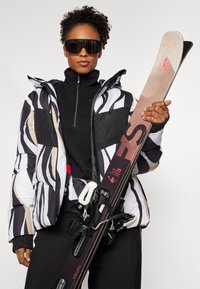 Pufferjakke med svart, hvit og beige zigzag-mønster, kombinert med en svart glidelås-topp og svarte bukser. Ski med bindinger i hånden.