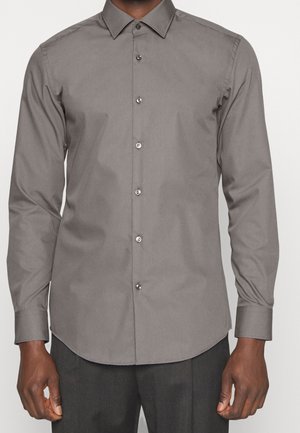 Mężczyzna w dopasowanej szarej koszuli na długi rękaw z kołnierzykiem w szpic i ciemnych spodniach, stojący na jednolitym tle.