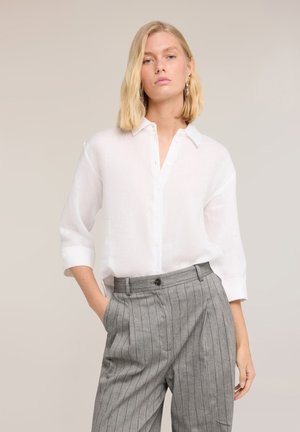 Blonde Frau trägt ein locker sitzendes weißes Hemd mit Dreiviertelärmeln und graue, hoch taillierte Nadelstreifenhose, Hand in der Tasche.