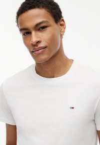 Jeune homme aux cheveux courts et bouclés portant un T-shirt blanc avec un petit logo rouge, blanc et bleu sur la poitrine, regardant la caméra.