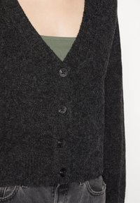 Czarny, teksturowany kardigan z głębokim dekoltem w kształcie litery V, z czterema czarnymi guzikami z przodu. Noszony na zielonej bluzce i szarych dżinsach.