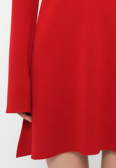 Close-up van een persoon die een rood gebreid jurkje met lange mouwen draagt, met een hand die gedeeltelijk zichtbaar is bij de zijsplit aan de zoom.