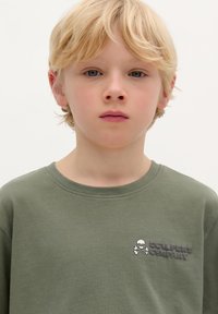 Olijfgroene katoenen T-shirt met een ronde hals, korte mouwen en een geborduurd logo met een doodskop en gekruiste botten op de borst.
