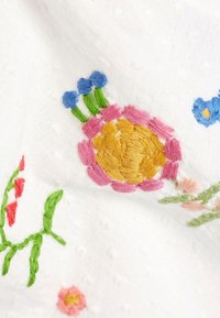 Witte stof met kleurrijke bloemenborduursels in roze, geel, blauw en groen. Heeft kenmerkende stikpatronen en een textuur oppervlak.