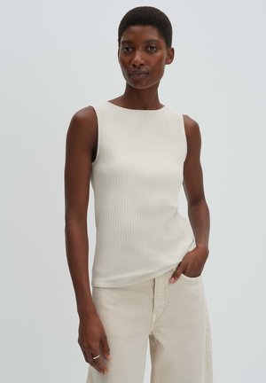 Frau mit kurzen Haaren, die ein ärmelloses, geripptes weißes Oberteil und hellfarbene Hosen trägt, steht mit einer Hand in der Tasche vor einem schlichten Hintergrund.