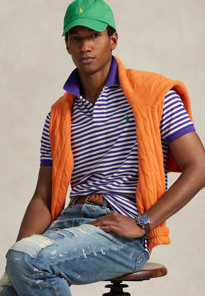 Άνδρας με πράσινο καπέλο, πόλο με μοβ και λευκές ρίγες, πορτοκαλί πουλόβερ περασμένο στους ώμους, τζιν με ξεβάμματα και καφέ ρολόι, καθισμένος σε σκαμπό.