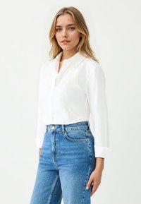 White button-up shirt made of a smooth fabric, featuring a classic collar, long sleeves, and rolled cuffs, paired with light blue high-waisted jeans.
