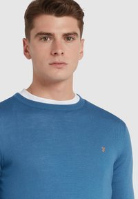 Niebieski sweter robiony na drutach z okrągłym dekoltem, z kontrastującym białym kołnierzykiem oraz małym pomarańczowym logo na piersi.