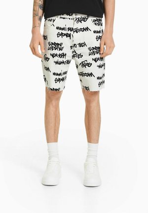 Mężczyzna w białych szortach z czarnym graffiti z nazwami miast, stojący na białym tle, widoczny od pasa do stóp.