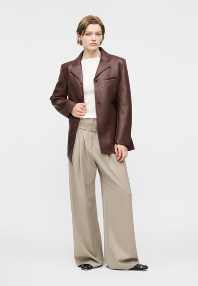 Γυναίκα που φοράει καφέ δερμάτινο σακάκι, λευκό πλεκτό πουλόβερ, μπεζ παντελόνι με φαρδιά μπατζάκια και ζώνη, και μαύρα παπούτσια, στέκεται μπροστά σε λευκό φόντο.