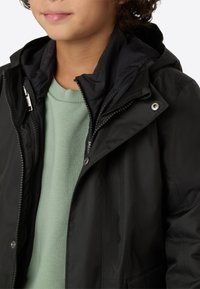 Černá bunda s kapucí, se zipem vpředu a sponkami. Je vrstvená přes světle zelený svetr s žebrovaným výstřihem.