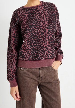 Personne portant un pull à manches longues bordeaux et noir imprimé léopard avec des poignets et un ourlet côtelés, associé à un pantalon marron taille haute.