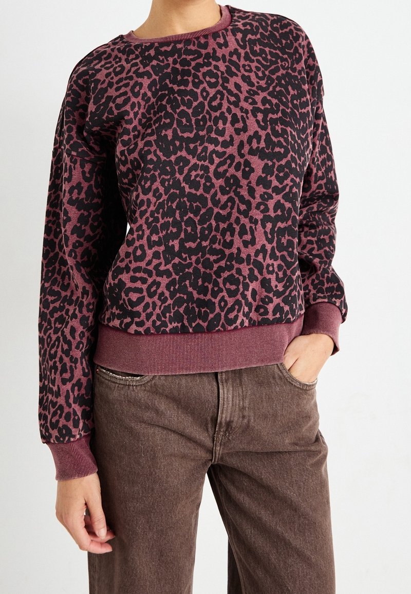 Personne portant un pull à manches longues bordeaux et noir imprimé léopard avec des poignets et un ourlet côtelés, associé à un pantalon marron taille haute.