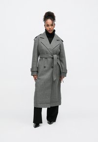 Harmaa villasekoitetrenssitakki, jossa on kaksiriviset napit, vyötärövyö ja olkatoppaukset. Yhdistetty mustaan poolopaitaan ja housuihin.