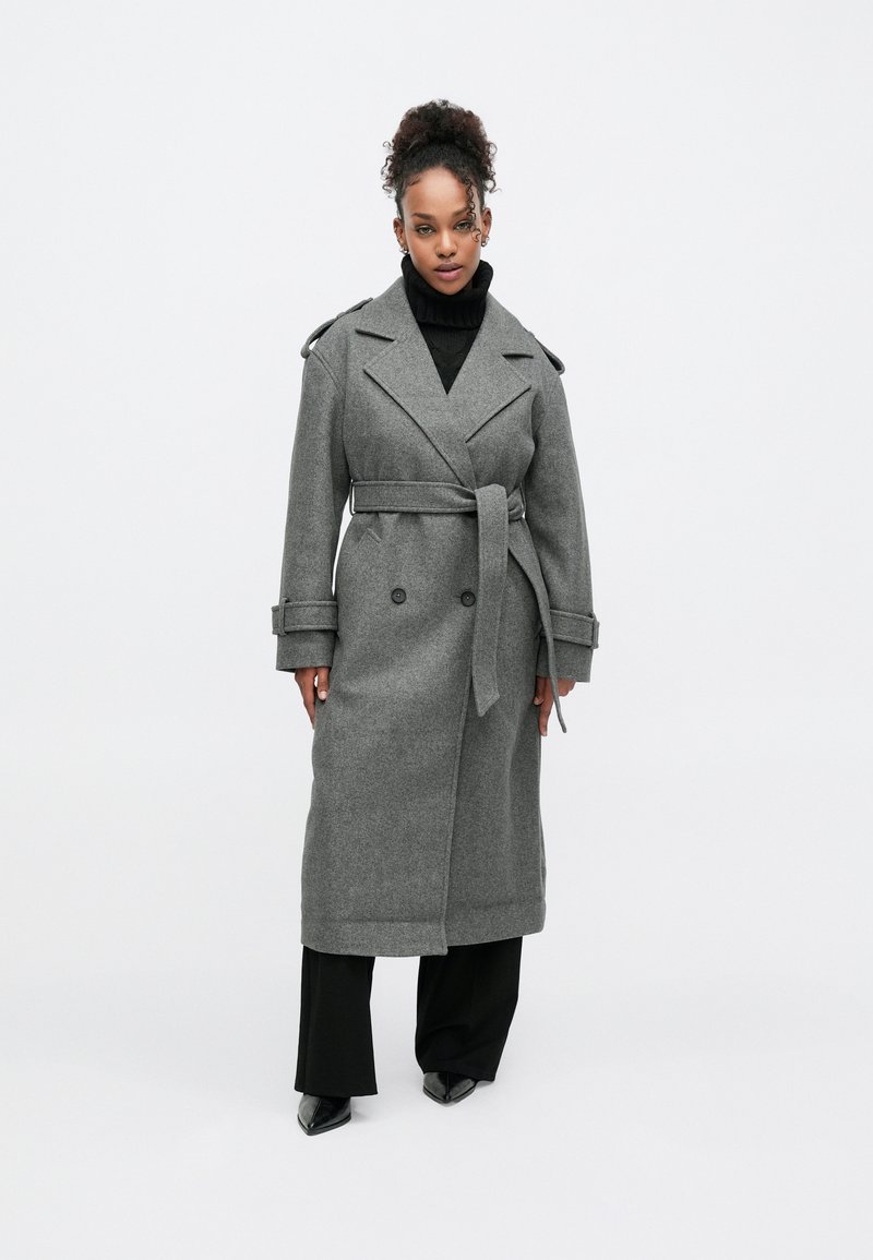 Harmaa villasekoitetrenssitakki, jossa on kaksiriviset napit, vyötärövyö ja olkatoppaukset. Yhdistetty mustaan poolopaitaan ja housuihin.