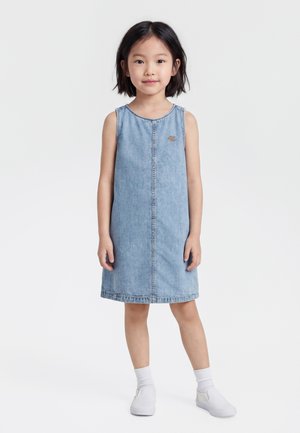 Nuori tyttö seisoo, hänellä on päällään hihaton vaaleansininen farkkumekko, valkoiset sukat ja valkoiset liukuvat kengät, yksinkertaista valkoista taustaa vasten.