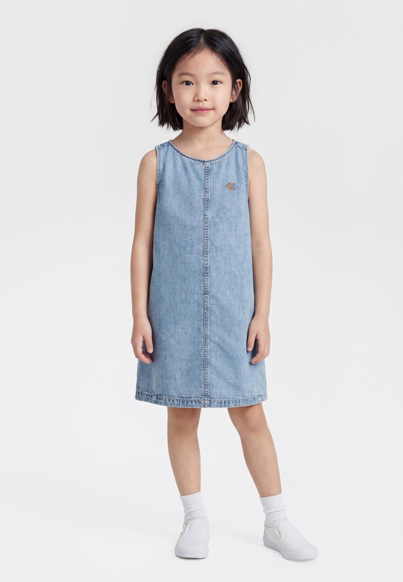 Νεαρή κοπέλα όρθια, ντυμένη με αμάνικο ανοιχτό μπλε φόρεμα από τζιν, λευκές κάλτσες και λευκά παπούτσια χωρίς κορδόνια, σε απλό λευκό φόντο.