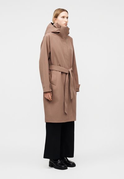 Γυναίκα που φοράει ένα μακρύ καφέ παλτό με ζώνη, ψηλό γιακά και κουκούλα, μαύρο παντελόνι με φαρδύ πόδι και μαύρα loafers, στέκεται μπροστά σε λευκό φόντο.