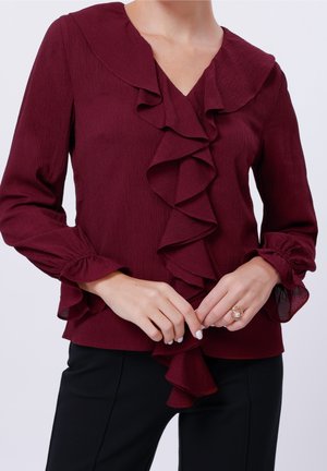 Femme portant un chemisier bordeaux à manches longues avec volants sur le devant et aux poignets, associé à un pantalon noir, mains jointes montrant une bague au doigt gauche.