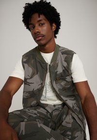 Chaleco de camuflaje con cuello verde acanalado, cierre de cremallera y bolsillo lateral, llevado sobre una camiseta gráfica ligera y pantalones cargo a juego.