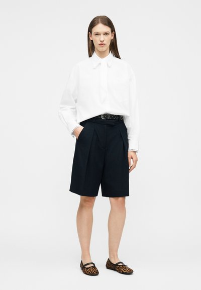 Femme debout portant une chemise blanche à manches longues, un short noir ceinturé à hauteur des genoux et des ballerines imprimé léopard sur un fond uni.