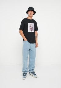 Svart t-skjorte med grafisk trykk, lyseblå løstsittende denim jeans, og en svart bøttehatt. Skoene er hvite med blå detaljer.
