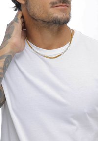 Silberne und goldene Kettenhalsketten übereinander getragen an einem nackten Hals, kombiniert mit einem schlichten weißen T-Shirt. Tätowierungen am Arm sichtbar.