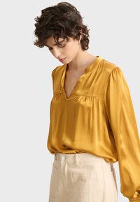 Mujer con cabello corto y rizado, vestida con una blusa amarilla mostaza de manga larga metida en unos pantalones beiges de tiro alto, mirando hacia abajo.