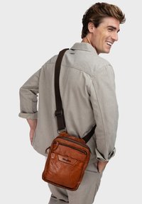 Bruine leren schoudertas met meerdere ritscompartimenten en een donkere verstelbare band, gedragen op de schouder. Gstructuursafwerking, compact ontwerp.