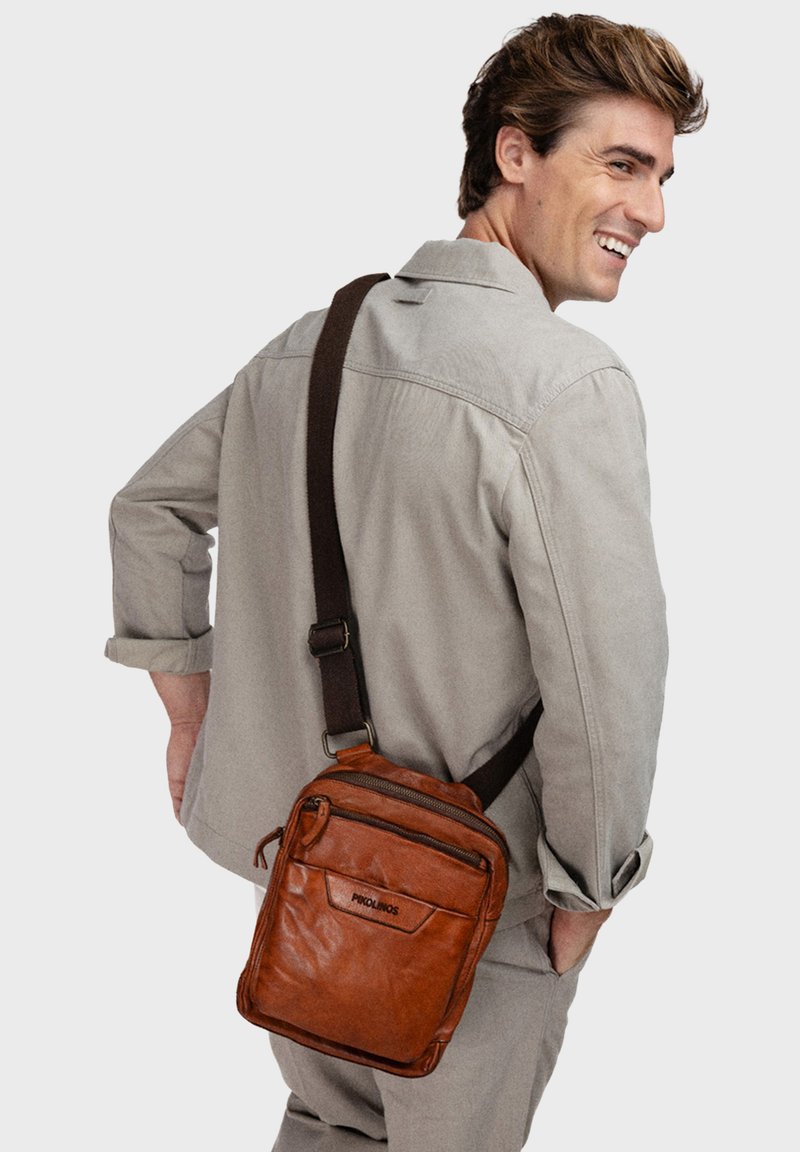 Bruine leren schoudertas met meerdere ritscompartimenten en een donkere verstelbare band, gedragen op de schouder. Gstructuursafwerking, compact ontwerp.