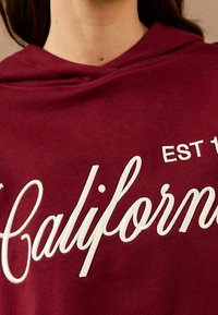 Bordeauxrode hoodie met witte geborduurde tekst "California," met "EST 19" erboven, gladde stof en een klassiek capuchonontwerp.