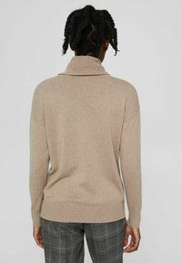 Pull en tricot beige avec un col haut, des épaules tombantes et des poignets côtelés. Texture douce, coupe décontractée, assorti à un pantalon à carreaux.