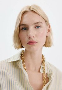 Blond, krótka fryzura typu bob; ubrana w jasnżółtą koszulę w niebieskie paski oraz masywny złoty łańcuch naszyjnika.