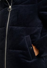 Närbild av en marinblå kordjacka med en silverdragkedja och en hand inskjuten i en knappslut ficka.