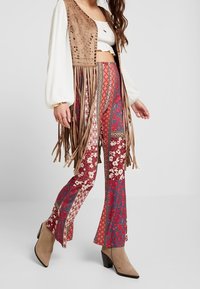 Fransiger brauner Wildleder-Westen über einem weißen Oberteil; farbenfrohe gemusterte Schlaghosen in Rot und Blau; beigefarbene Blockabsatz-Stiefeletten.