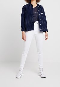 Chaqueta a rayas en azul marino de tejido grueso, con cierre de botones, combinada con jeans blancos ajustados. Modelo usando zapatillas altas blancas.