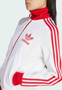 Bijela jakna na zip s visokim ovratnikom, crvenim detaljima i tri crvene pruge na rukavima. Sadrži mali crveni Adidas logotip na prsima.