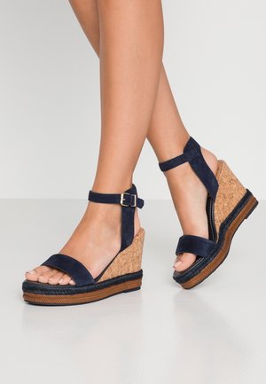 Granatowe zamszowe sandały na koturnie z korkowym obcasem, paskiem wokół kostki oraz szerokim paskiem na palce; teksturowana gumowa podeszwa z plecionym akcentem.