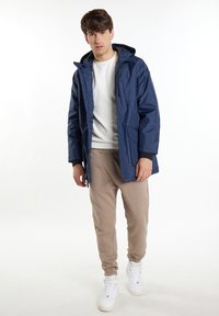 Marinblå vattentät jacka med huva, med dragkedja fram och sidofickor, bärs över en vit skjorta och beigea sweatpants, kompletterad med vita sneakers.