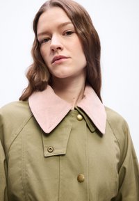 Νεαρή γυναίκα με κυματιστά καστανά μαλλιά, που φοράει ένα ελαιόχρωμο παλτό με μεγάλο ροζ γιακά από κορντούρο και ορειχάλκινες κουμπότρυπες, με ουδέτερη έκφραση.