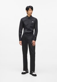 Melnas pogcaurules krekls ar spīdīgu apkakli, kopā ar pielāgotām melnām biksēm un melnām kurpēm ar baltām akcentiem. Ievērojams logotipa elements uz kreklā.