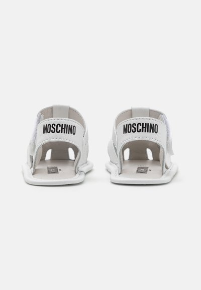 Białe sandały z wycinanym wzorem. Posiadają czarne logo "MOSCHINO" na pięcie. Wykonane z miękkiego materiału, z płaską podeszwą i minimalistycznym stylem.