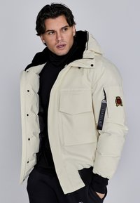 Giacca invernale imbottita di colore crema con cappuccio removibile, tasche con zip e logo ricamato sulla manica. Fodera interna nera.